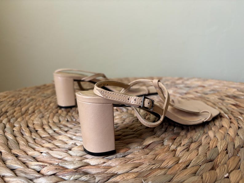 May include: A pair of beige sandals with a block heel and a thin ankle strap featuring a small metal buckle. The sandals have a square toe design and a smooth finish, with the heel and straps constructed in a matching light tan color.