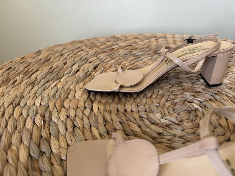 May include: A pair of beige sandals with a square toe, a circular accent on the front strap, a thin T-strap design, and a block heel. The sandals feature a delicate ankle strap with a small buckle closure and a smooth, neutral-toned finish.