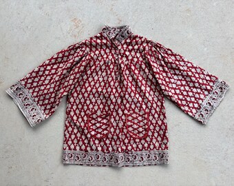 May include: A long-sleeved shirt featuring a deep red and white repeating floral block print pattern. The garment includes a high collar, two front patch pockets, and wide sleeves with a detailed decorative border along the cuffs and hem.