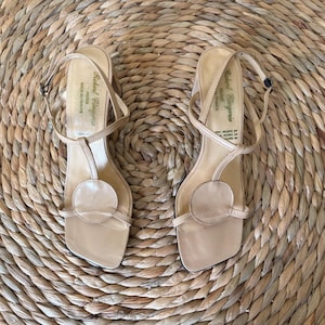 May include: A pair of beige slingback sandals with a square toe, a T-strap design, and a circular accent on the vamp. The insoles feature the text Robert Clergerie Paris Made in France. The sandals have a thin ankle strap with a small buckle.