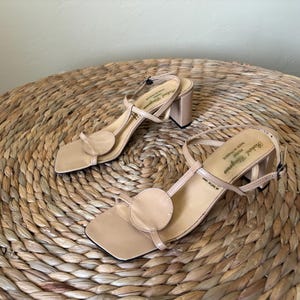May include: A pair of beige sandals with a square toe, a circular accent on the front strap, and a block heel. The slingback strap features a buckle closure, and the insole is printed with the text Robert Clergerie Paris Made in France.