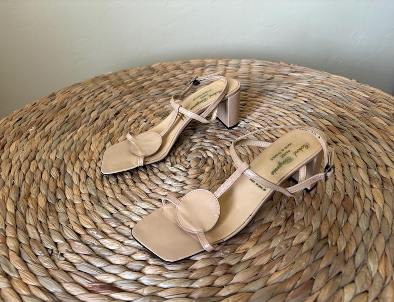 May include: A pair of beige sandals with a square toe, a circular accent on the front strap, and a block heel. The slingback strap features a buckle closure, and the insole is printed with the text Robert Clergerie Paris Made in France.