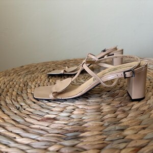 May include: A pair of beige sandals with a square toe, a block heel, and thin straps that form a T-strap design. The sandals feature a small adjustable buckle on the side strap and a smooth finish, providing a clean and structured look for footwear.