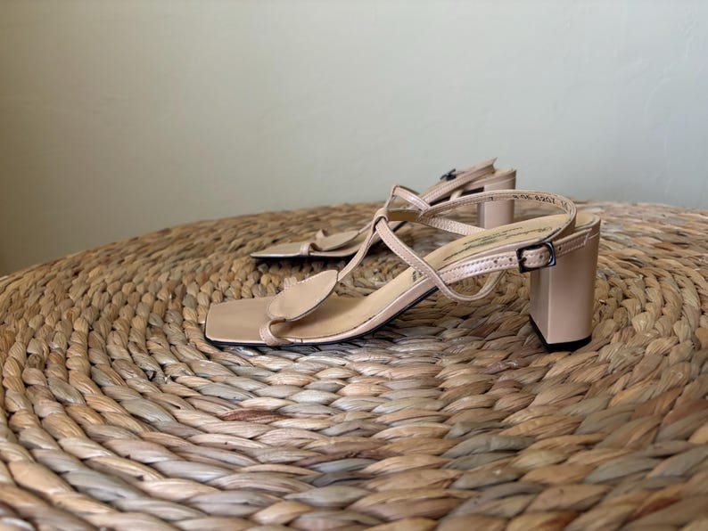 May include: A pair of beige sandals with a square toe, a block heel, and thin straps that form a T-strap design. The sandals feature a small adjustable buckle on the side strap and a smooth finish, providing a clean and structured look for footwear.