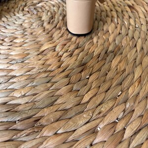 May include: A round, woven natural fiber mat with a braided texture in shades of tan and light brown. Two cylindrical, light beige furniture legs with black circular bases are positioned on the surface of the mat, showing the base of the object.
