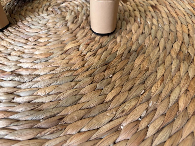 May include: A round, woven natural fiber mat with a braided texture in shades of tan and light brown. Two cylindrical, light beige furniture legs with black circular bases are positioned on the surface of the mat, showing the base of the object.