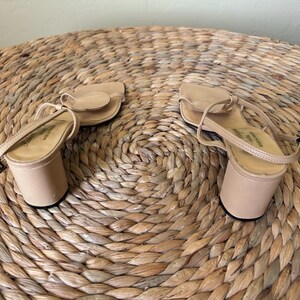 May include: A pair of beige sandals with a block heel, a thin ankle strap, and a rounded toe strap featuring a circular accent. The sandals have a smooth finish and a simple construction, showcasing a neutral color palette and a sturdy heel design.