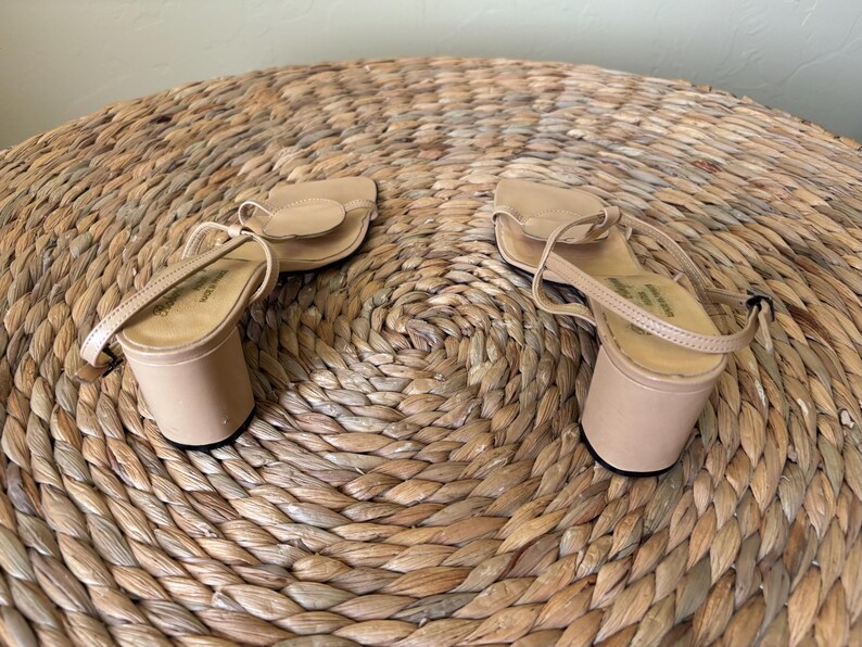 May include: A pair of beige sandals with a block heel, a thin ankle strap, and a rounded toe strap featuring a circular accent. The sandals have a smooth finish and a simple construction, showcasing a neutral color palette and a sturdy heel design.