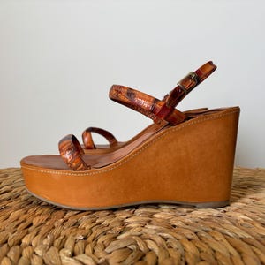 Puede incluir: Sandalias de cuña con plataforma de cuero marrón. Las sandalias presentan una suela de plataforma, un tacón de cuña y correas con estampado de reptil. Cierre de hebilla. Sobre una superficie tejida.