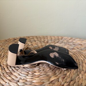May include: A pair of beige sandals with a block heel and a black sole. The sandals feature thin straps and a square toe design. The black soles show signs of wear with visible scuffing and worn areas on the bottom surface of the footwear.