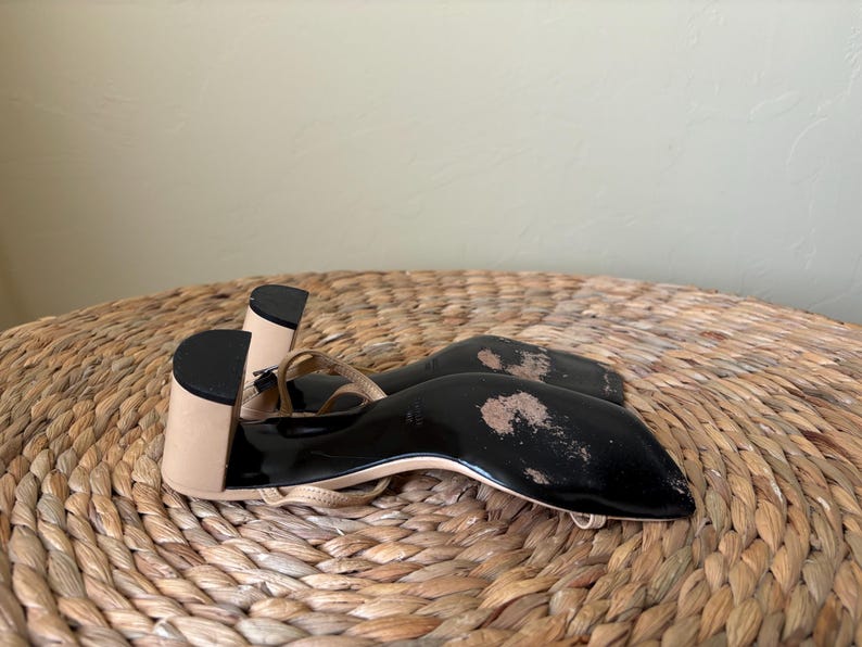 May include: A pair of beige sandals with a block heel and a black sole. The sandals feature thin straps and a square toe design. The black soles show signs of wear with visible scuffing and worn areas on the bottom surface of the footwear.