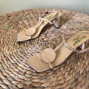 May include: A pair of beige leather sandals with a square toe, a circular accent on the front strap, a thin T-strap design, and a block heel. The interior insole features the text Robert Clergerie Paris Made in France.