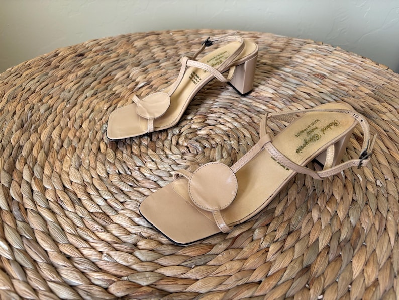 May include: A pair of beige leather sandals with a square toe, a circular accent on the front strap, a thin T-strap design, and a block heel. The interior insole features the text Robert Clergerie Paris Made in France.