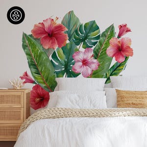 May include: A wall decal featuring a watercolor design of tropical flowers and leaves in shades of pink, red, and green. The decal includes hibiscus flowers and large, lush leaves, creating a vibrant, botanical theme. The decal is placed above a bed with white bedding.