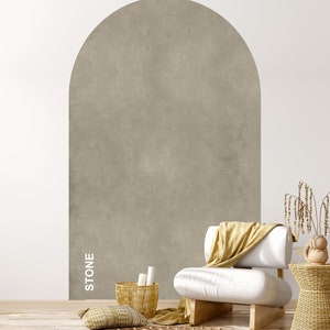 Op de afbeelding: Een witte fauteuil met een gele deken staat voor een muur met een groot, gewelfd, getextureerd, grijs stenen paneel. De tekst "STONE" staat verticaal op het paneel. Een geweven mand met witte rollen staat op de vloer naast de fauteuil.