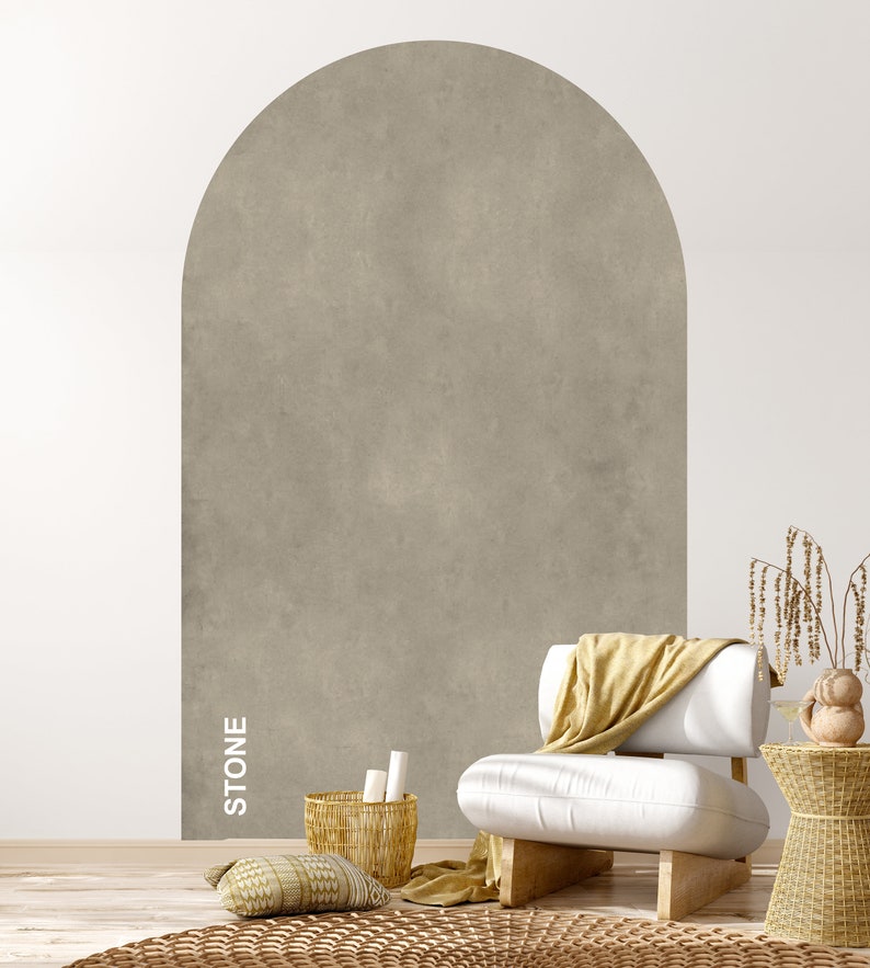 Op de afbeelding: Een witte fauteuil met een gele deken staat voor een muur met een groot, gewelfd, getextureerd, grijs stenen paneel. De tekst "STONE" staat verticaal op het paneel. Een geweven mand met witte rollen staat op de vloer naast de fauteuil.