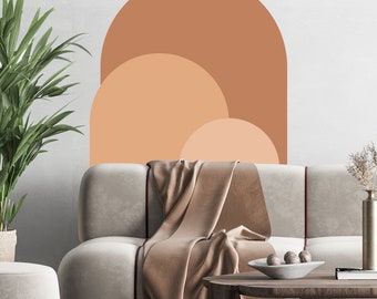 Puede incluir: Una calcomanía de pared abstracta de color marrón claro, beige y melocotón con forma de arco redondeado. La calcomanía está en una pared blanca detrás de un sofá marrón claro con una manta de lana marrón.
