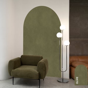 May include: A green velvet armchair sits in front of a green wall with an arched cutout. A chrome floor lamp with three white globe lights stands next to a brown leather sofa. The wall behind the sofa has a green swatch with the word "OLIVE" written on it.