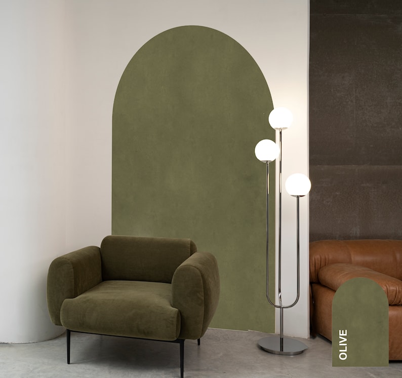 Op de afbeelding: Een groene fluwelen fauteuil staat voor een groene muur met een boogvormige uitsparing. Een chromen staande lamp met drie witte bolvormige lampen staat naast een bruine leren bank. De muur achter de bank heeft een groene kleurstaal met het woord "OLIVE" erop.