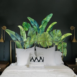 May include: A white bed with a gold metal headboard and a tropical leaf patterned wall decal. The bed has white bedding and a black and white patterned pillow.