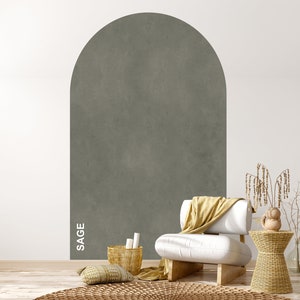 Op de afbeelding: Een witte fauteuil met een gele deken staat voor een muur met een boog in saliegroen. Een geweven mand met kaarsen staat op de vloer naast een geweven bijzettafel met een klein decoratief object erop.