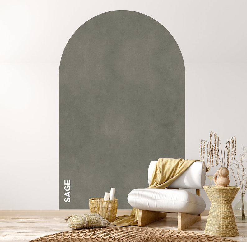 Op de afbeelding: Een witte fauteuil met een gele deken staat voor een muur met een boog in saliegroen. Een geweven mand met kaarsen staat op de vloer naast een geweven bijzettafel met een klein decoratief object erop.