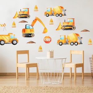 May include: A set of yellow and orange construction vehicle wall decals, including a dump truck, cement mixer, excavator, and bulldozer. The decals are on a white wall in a child's room with a table and chairs.