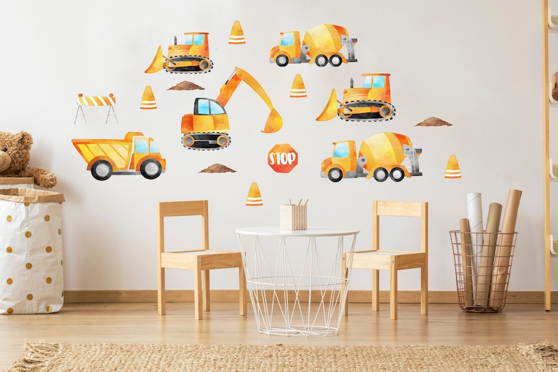 May include: A set of yellow and orange construction vehicle wall decals, including a dump truck, cement mixer, excavator, and bulldozer. The decals are on a white wall in a child's room with a table and chairs.