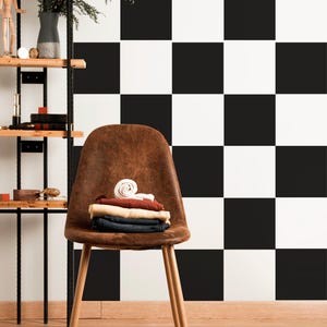 May include: A brown leather chair with wooden legs holds folded clothes. A pair of black dress shoes sits on a rug in front of the chair. The background features a black and white chequered wall and a wooden shelf with decorative items.
