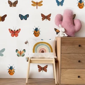 May include: A white wall with a variety of colorful butterfly and ladybug wall decals. The decals are arranged in a random pattern. There is a wooden dresser with three drawers and a pink cactus pillow on top of the dresser. There is also a wooden stool with a white pillow with a rainbow design on it.