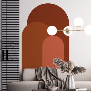 May include: A living room interior with a white wall featuring a large abstract wall decal of three overlapping arches in shades of brown and orange. A gray couch with a gray knitted throw blanket is positioned in front of the wall. A modern light fixture with round white globes hangs from the ceiling.
