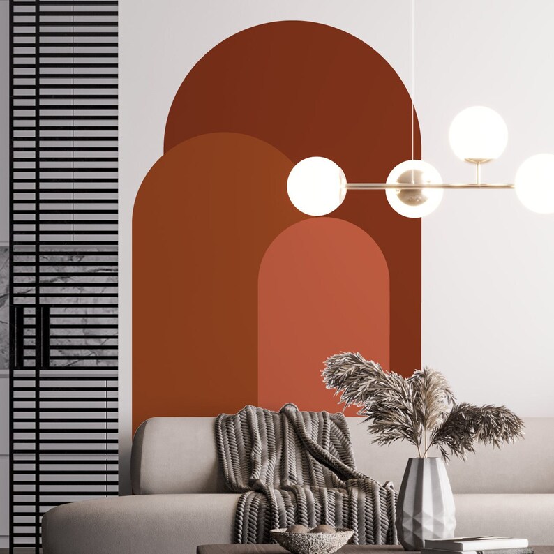 May include: A living room interior with a white wall featuring a large abstract wall decal of three overlapping arches in shades of brown and orange. A gray couch with a gray knitted throw blanket is positioned in front of the wall. A modern light fixture with round white globes hangs from the ceiling.