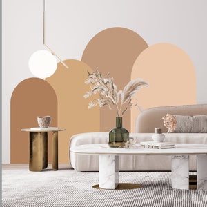 May include: A living room interior design with a white couch, a white and gold coffee table, and a gold side table. The wall is painted with a light brown and peach color scheme with a geometric arch design. A vase with dried flowers sits on the coffee table.