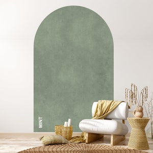 Op de afbeelding: Een witte fauteuil met een gele deken staat voor een muur met een grote, gebogen, mintgroene accentmuur. De muur heeft een textuur, geschilderde afwerking. Een kleine, geweven bijzettafel met een decoratief object staat naast de fauteuil.