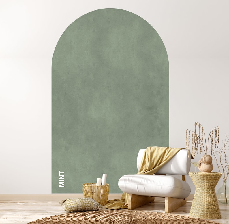 Op de afbeelding: Een witte fauteuil met een gele deken staat voor een muur met een grote, gebogen, mintgroene accentmuur. De muur heeft een textuur, geschilderde afwerking. Een kleine, geweven bijzettafel met een decoratief object staat naast de fauteuil.