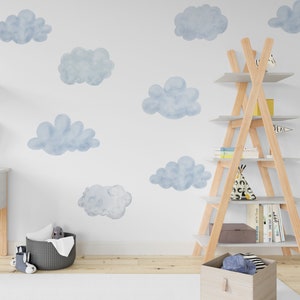 Puede incluir: Una pared blanca con calcomanías de nubes de acuarela azul. Una estantería de madera en forma de escalera con libros y una pequeña tienda de campaña tipi en los estantes. Una cesta tejida gris con un animal de peluche en la parte superior.