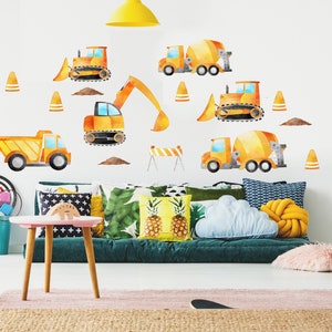 May include: A yellow and orange construction vehicle wall decal set with a dump truck, excavator, cement mixer, and bulldozer. The decals are on a white wall in a child's room with a green couch and pillows.