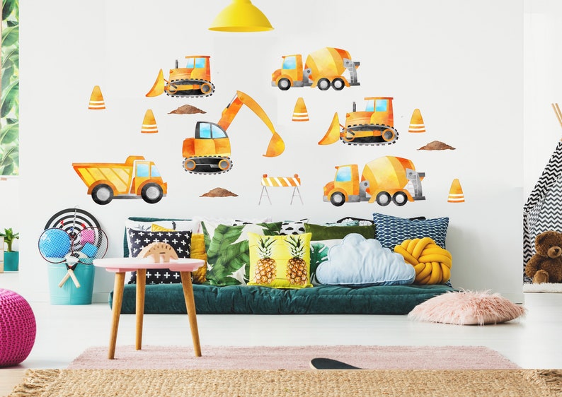 May include: A yellow and orange construction vehicle wall decal set with a dump truck, excavator, cement mixer, and bulldozer. The decals are on a white wall in a child's room with a green couch and pillows.