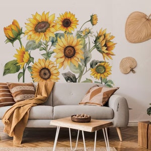 May include: A grey sofa with brown throw cushions and a blanket sits in front of a wall with a sunflower decal. A wooden coffee table with a bowl of nuts sits in front of the sofa.