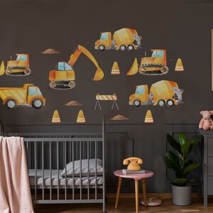 May include: A nursery room with a gray crib and a dresser with gold drawer pulls. The walls are decorated with yellow construction vehicle decals, including a cement mixer, a bulldozer, and an excavator. There are also orange traffic cones and a barricade.