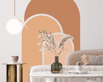 Puede incluir: Un sofá blanco con un cojín beige se encuentra frente a una pared con un gran diseño de arco abstracto en tonos naranja y melocotón. Un jarrón con flores secas y una mesa de café con un libro y un pequeño objeto decorativo están en primer plano.
