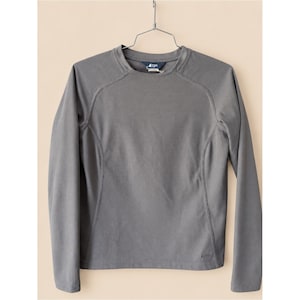 May include: A long-sleeve crew neck shirt in a solid medium gray color. The garment features raglan-style seams extending from the neckline to the underarm, a straight hemline, and a small logo detail near the bottom right corner on the front.