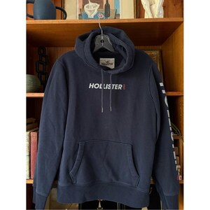 May include: A navy blue hooded sweatshirt featuring a front kangaroo pocket and long sleeves. The word HOLLISTER is printed in white on the chest with a small red USA text, and the brand name is printed vertically in white down the left sleeve.