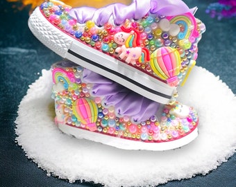 Puede incluir: Un par de zapatos de lona blancos decorados con brillantes, cuentas y abalorios de colores, incluyendo un unicornio, un arcoíris y un globo aerostático. Los zapatos tienen cintas de satén morado.