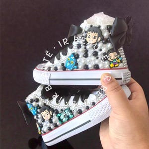 May include: White high-top sneakers embellished with black and white beads, cartoon characters, and black bows. The sneakers have a white rubber sole and are decorated with rhinestones. The text "Té • IR BE" is visible on the side.