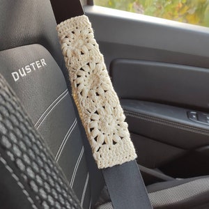 Puede incluir: Funda de cintur&oacute;n de seguridad de ganchillo color crema con un patr&oacute;n floral. La funda est&aacute; envuelta alrededor de un cintur&oacute;n de seguridad negro, con un asiento de coche negro visible en el fondo. La palabra "DUSTER" est&aacute; bordada en el asiento.