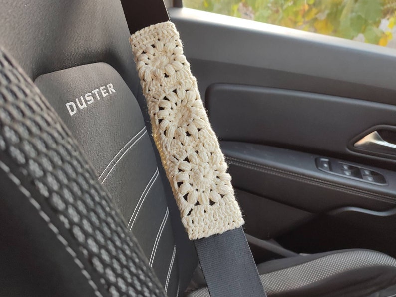 Puede incluir: Funda de cintur&oacute;n de seguridad de ganchillo color crema con un patr&oacute;n floral. La funda est&aacute; envuelta alrededor de un cintur&oacute;n de seguridad negro, con un asiento de coche negro visible en el fondo. La palabra "DUSTER" est&aacute; bordada en el asiento.