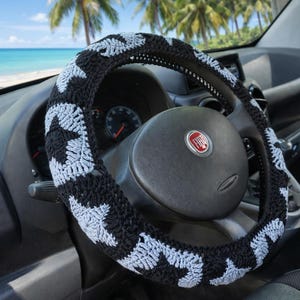 A black and grey steering wheel cover with a star pattern. The cover is on a black steering wheel inside a car. The background shows a beach scene with palm trees and blue water.