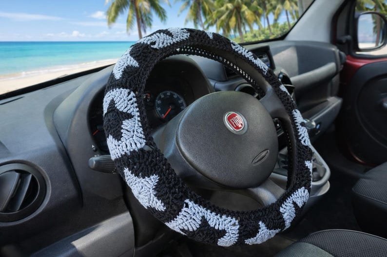 A black and grey steering wheel cover with a star pattern. The cover is on a black steering wheel inside a car. The background shows a beach scene with palm trees and blue water.