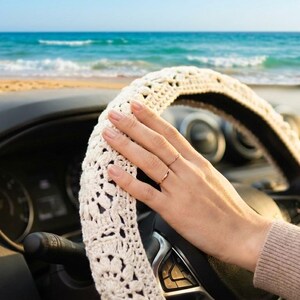 A ivory steering wheel cover is shown in a car with a beach and ocean in the background. The cover has an open, lacy design. The car's dashboard and steering wheel are black.